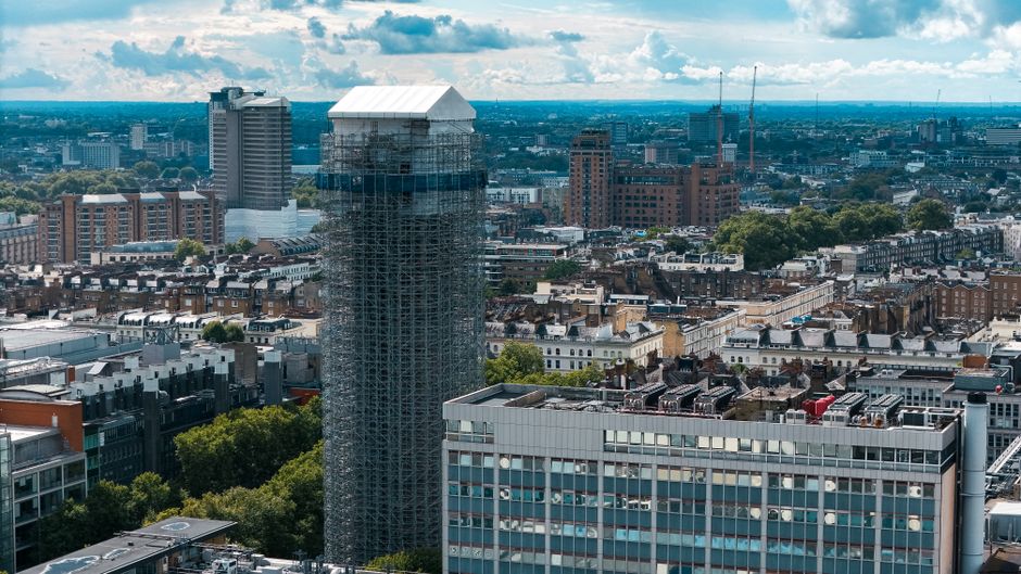 An aerial view of a cityscape featuring a high-rise building under construction, covered in scaffolding and wrapped in protective sheeting. Surrounding the construction site are various office and res